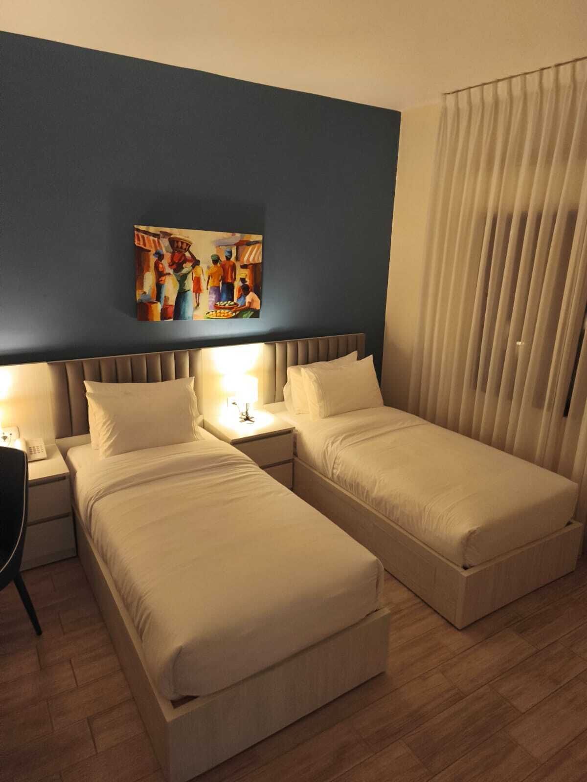 A hotel room with two single beds, a nightstand with a lamp, a painting on the wall, and a window with curtains.