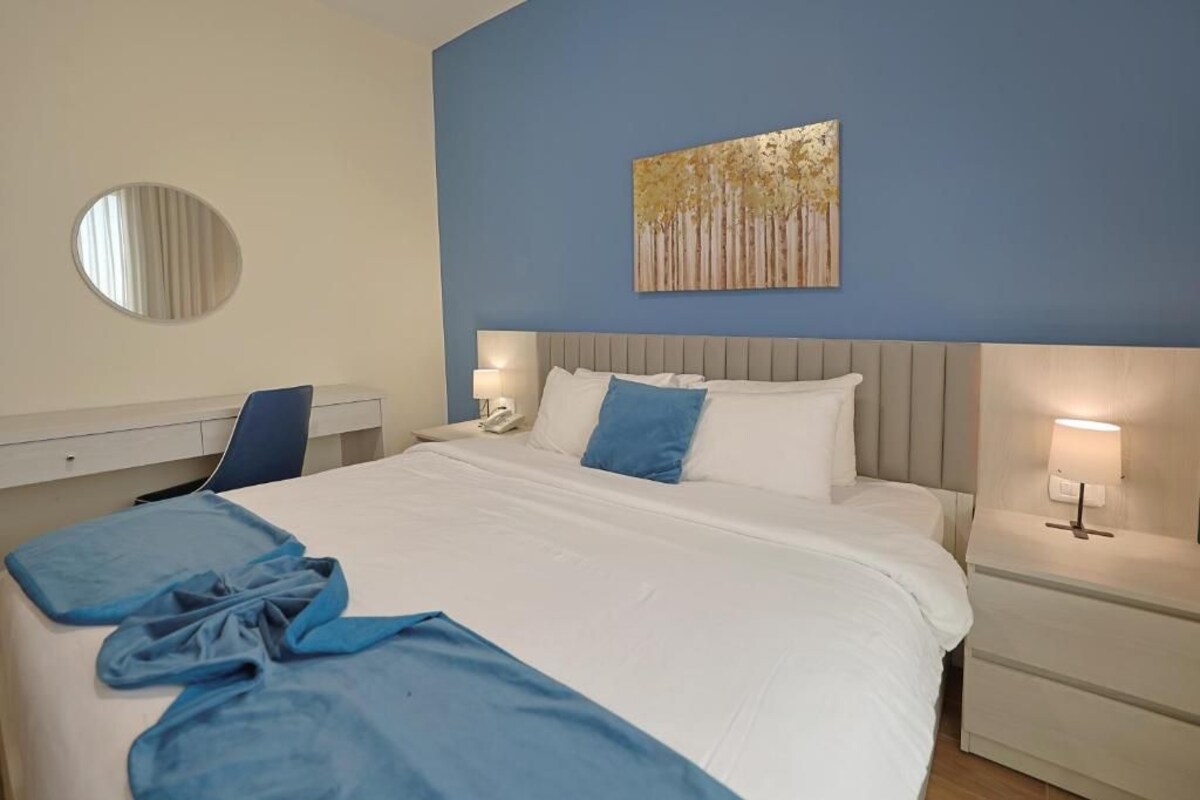 A hotel room with a bed, a desk, a chair, a bedside table with a lamp, and a wall-mounted artwork.