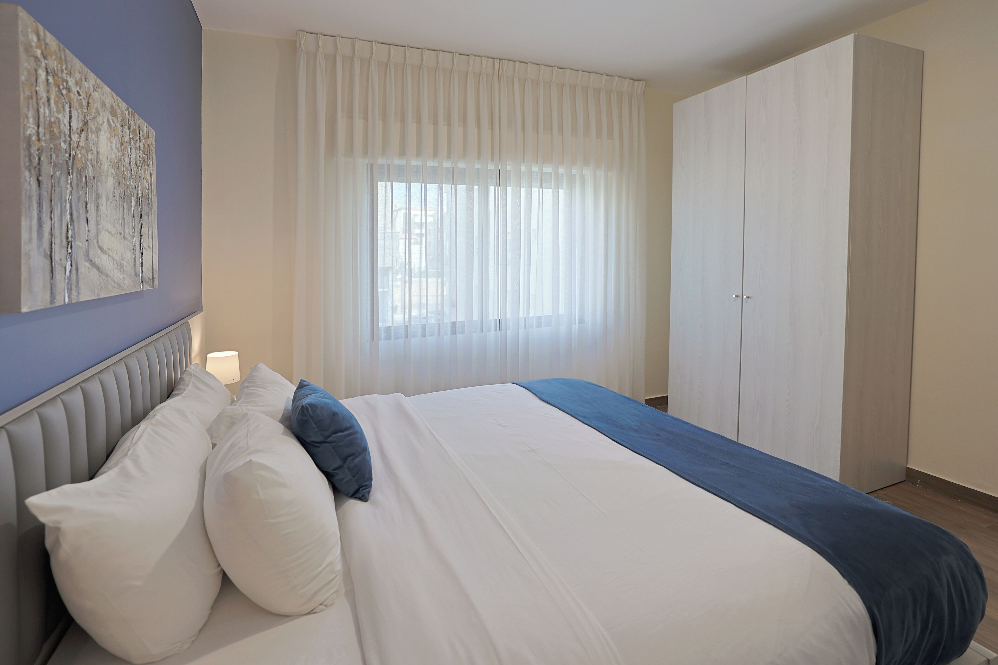 Un dormitorio con cama, mesita de noche, ventana con cortinas, armario y una obra de arte colgada en la pared.