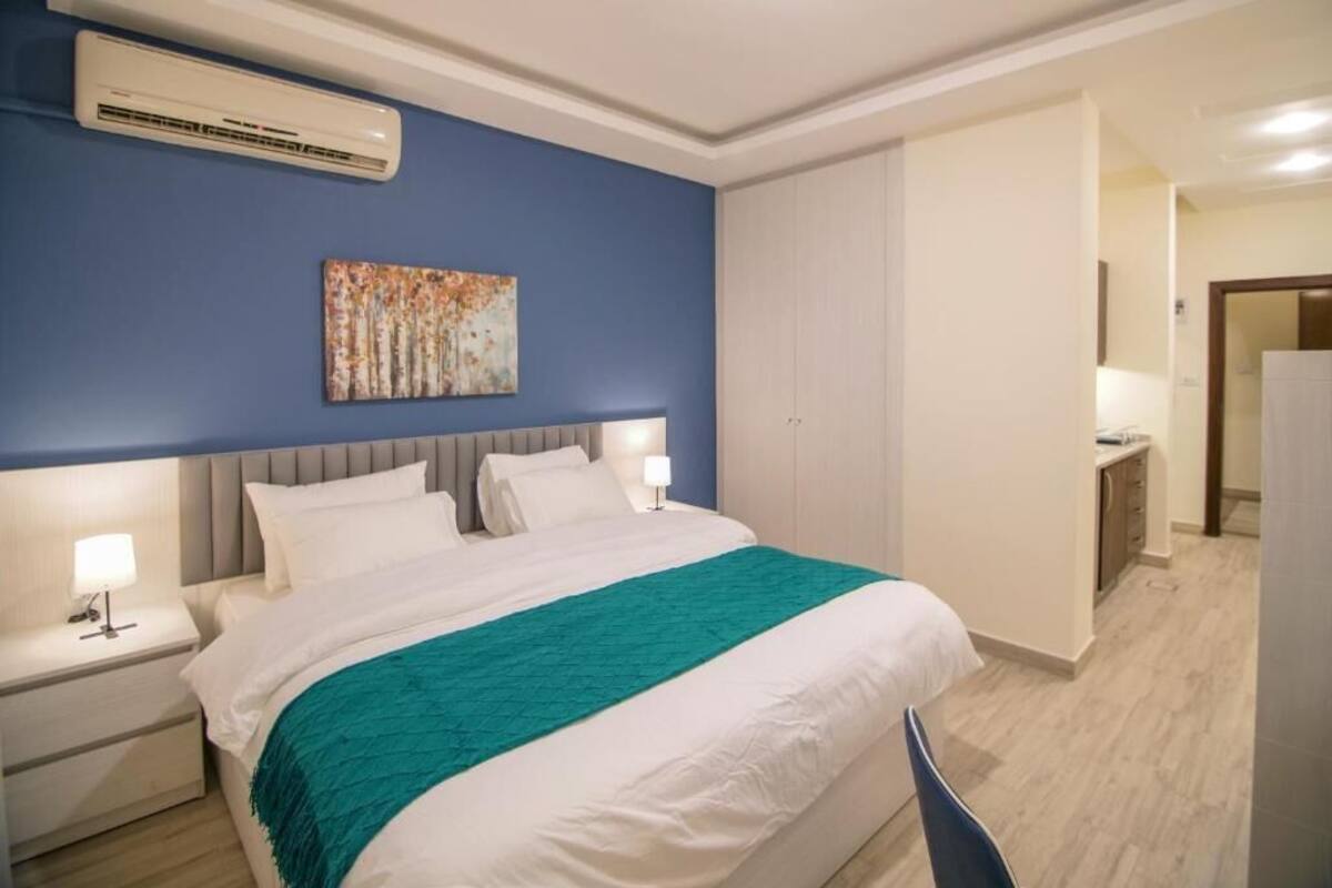 A hotel room with a bed, bedside tables, a nightstand, a wall-mounted air conditioner, a painting, and a wardrobe.