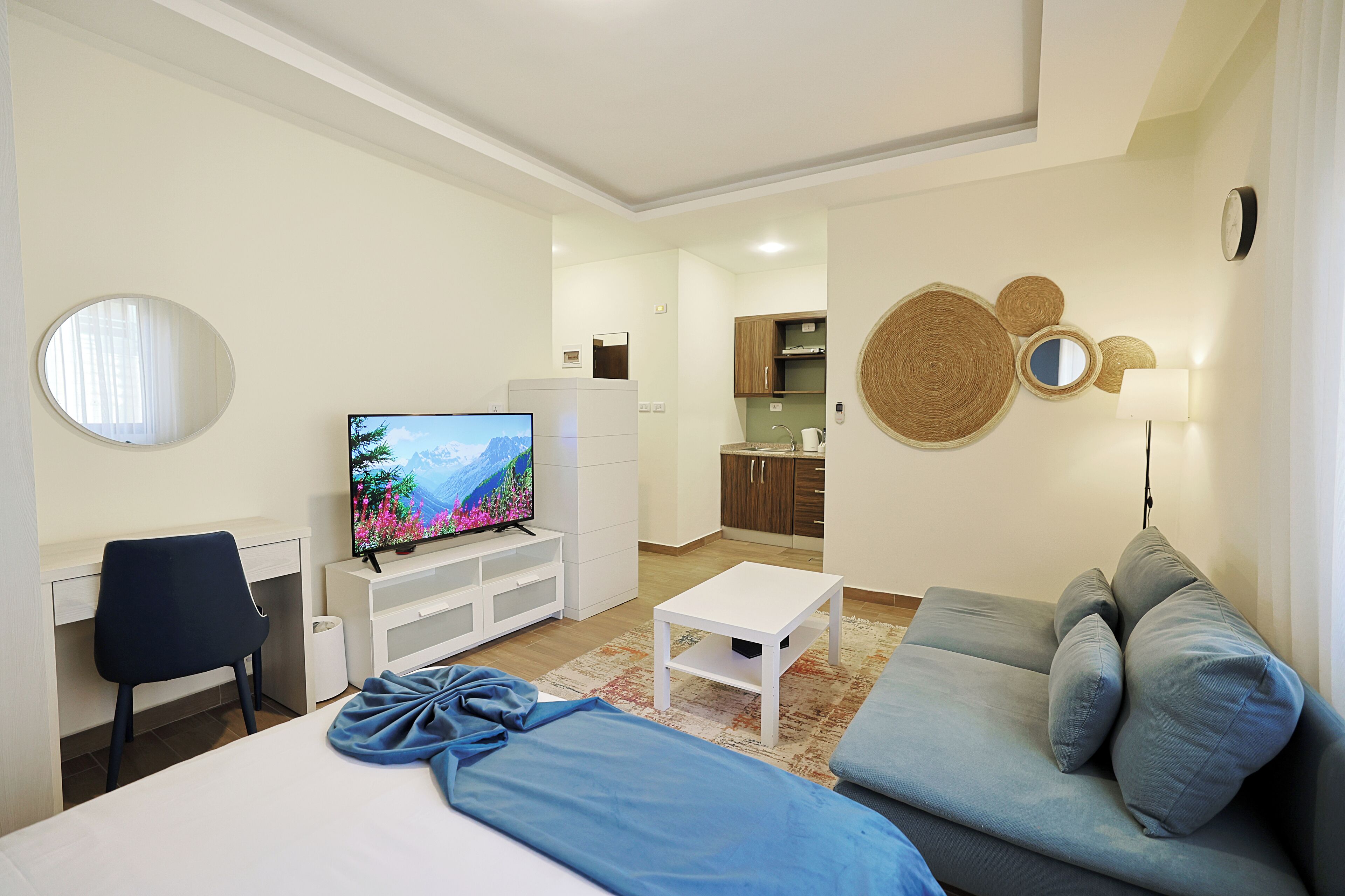 A modern hotel room with a bed, a TV, a sofa, a desk, and a kitchen area in the background.