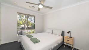 Un dormitorio con cama, mesita de noche, lámpara de escritorio, ventilador de techo y ventana con vista a follaje.