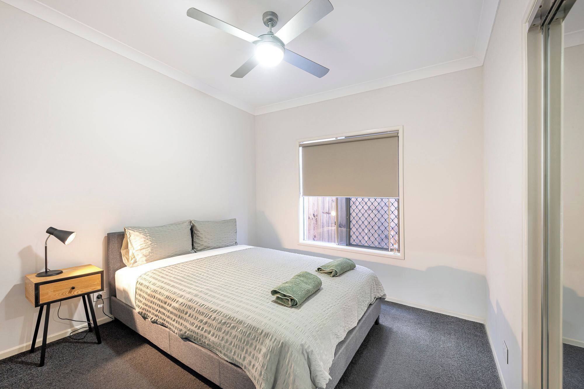A bedroom with a bed, a nightstand, a ceiling fan, a window with a blind, and a door.