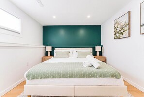A neatly made bed with a green and white striped bedspread, a white pillow, and a rolled white towel. There are two bedside tables with lamps, a dark green headboard, and a framed picture on the wall.