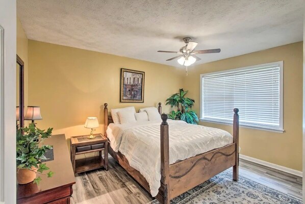 A bedroom with a wooden four-poster bed, a ceiling fan, a nightstand with a lamp, a potted plant, and a window with blinds.
