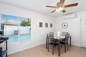 A dining area with a table and chairs, a ceiling fan, a clock, and a window with blinds.