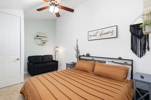 A bedroom with a bed, a ceiling fan, a wall-mounted sign, a black sofa, a nightstand, and a macrame wall hanging. - Extravagant Phoenix Retreat|Pool|Gazebo|HeatedPool (Phoenix)