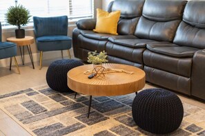 A modern living room with a dark leather sofa, a round wooden coffee table, and two black poufs.