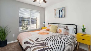 Une chambre à coucher comprenant un lit, un ventilateur de plafond, une fenêtre avec des stores, une table de chevet avec une lampe et une œuvre d’art accrochée au mur.