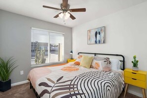 A bedroom with a bed, a ceiling fan, a window with blinds, a bedside table with a lamp, and a wall-mounted artwork.