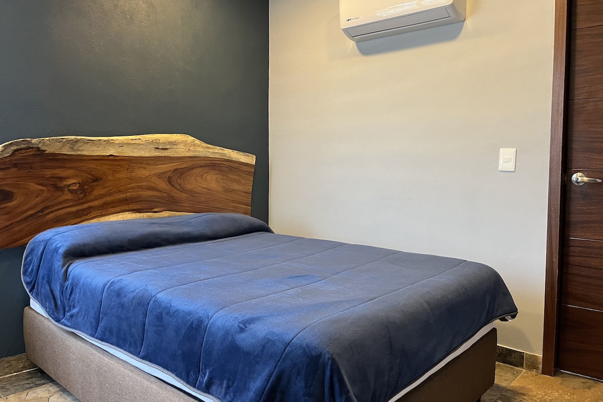 A bedroom with a wooden headboard, a bed with a blue bedspread, an air conditioning unit on the wall, and a wooden door.