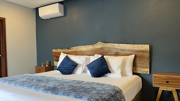 A bedroom with a wooden headboard, a bed with white and blue bedding, a bedside table with a lamp, and a wall-mounted air conditioner. - Hotel La Bikina (Tequila)