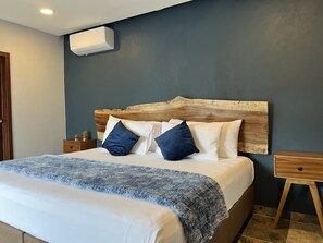 A bedroom with a wooden headboard, a bed with white and blue bedding, a bedside table with a lamp, and a wall-mounted air conditioner.