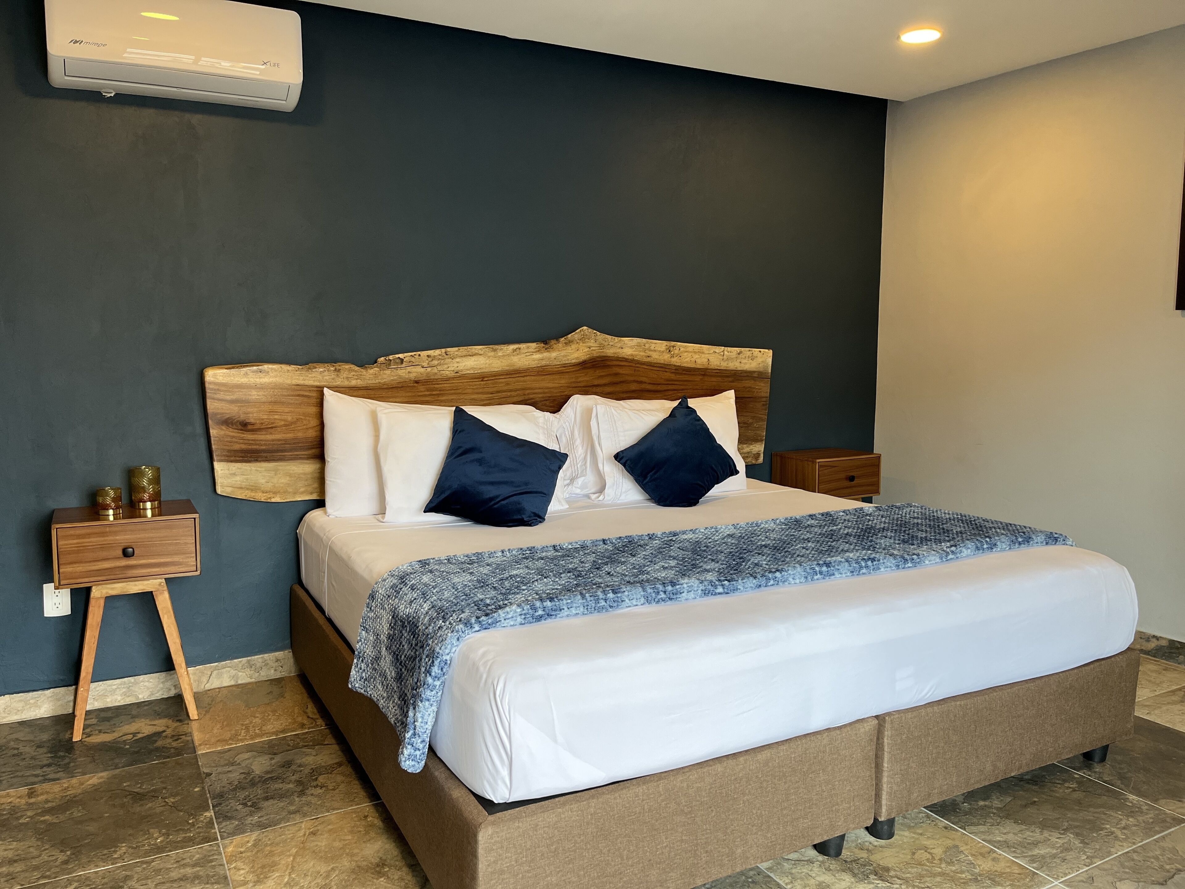 A bedroom with a wooden headboard, a bed with white and dark blue pillows, a blue knitted throw, and a nightstand with a wooden drawer and two decorative vases.