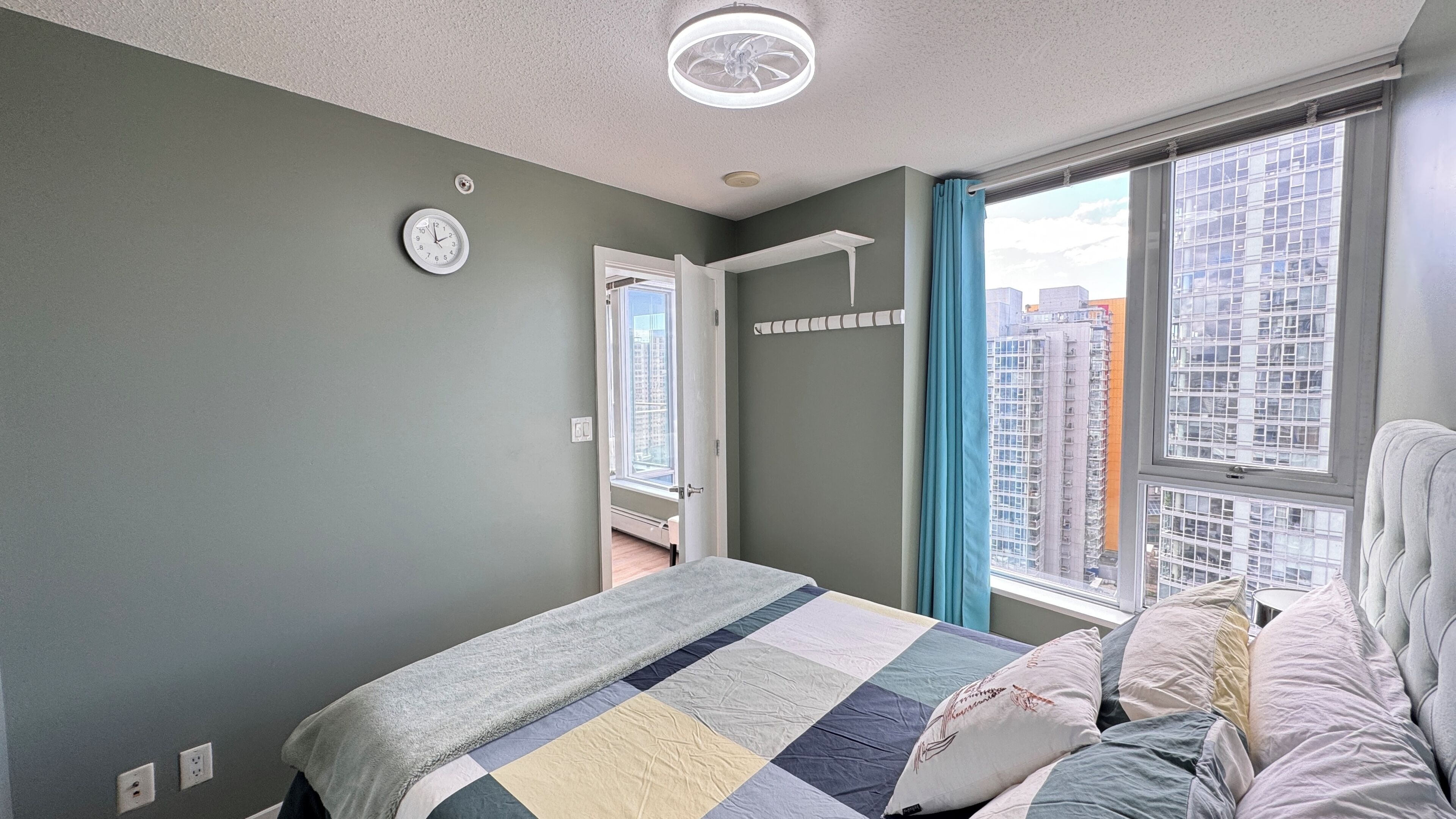 A bedroom with a bed, a window with curtains, a wall clock, and a door leading to another room.