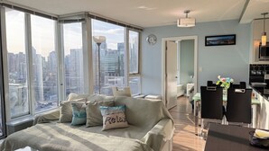 A modern living room with a large window, a sofa with cushions, a dining area with a table and chairs, and a kitchen area. - Downtown High-rise Condo Amazing View+Pool+Hot Jacuzzi+Sauna+Gym+ free Parking (Vancouver)