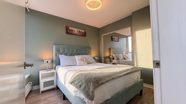 A bedroom with a tufted headboard, a bed with white bedding, two bedside tables with lamps, a painting on the wall, and a mirror reflecting the room.