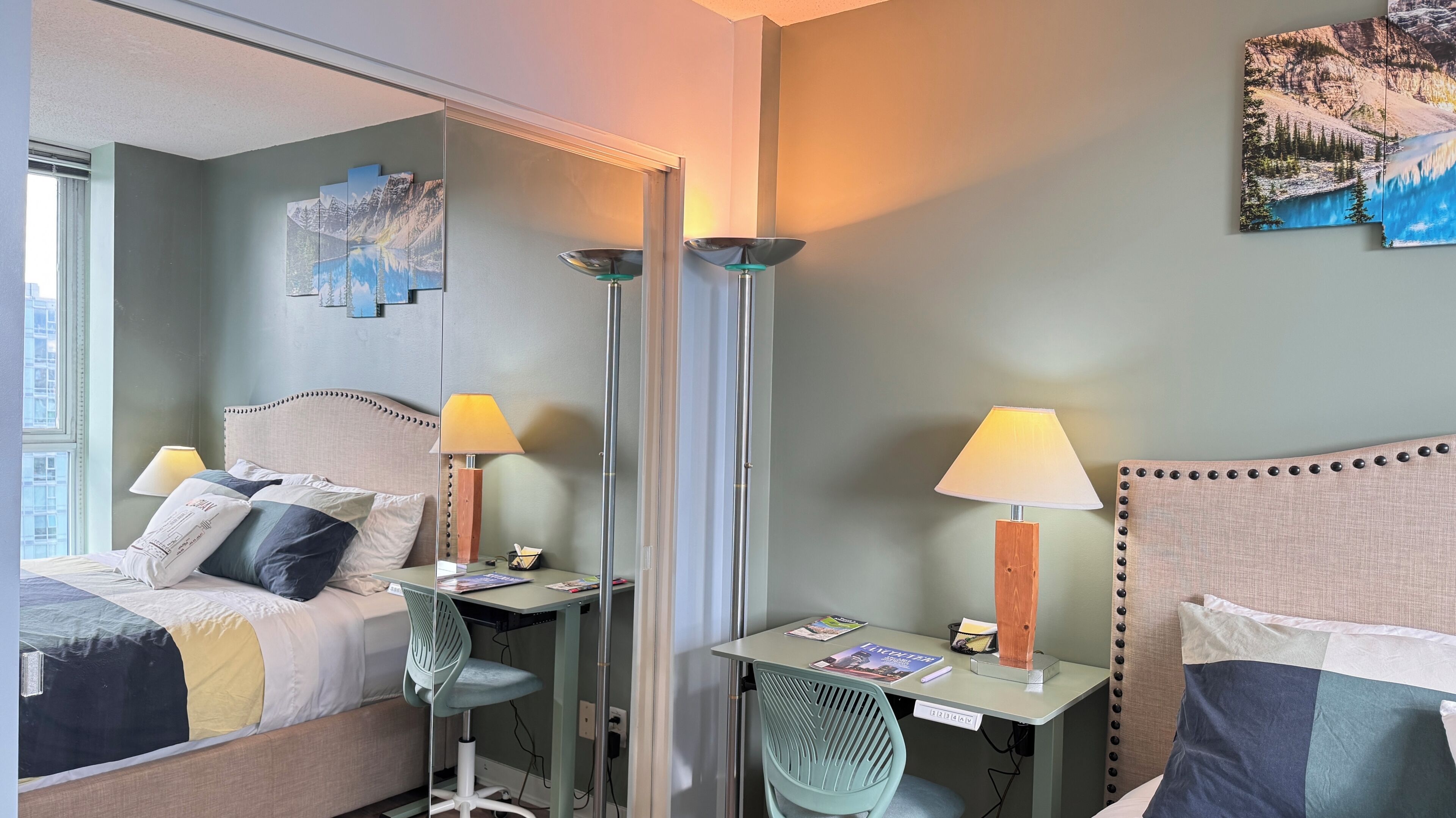 A hotel room with a bed, a desk, a chair, two lamps, a mirror, and artwork on the walls.