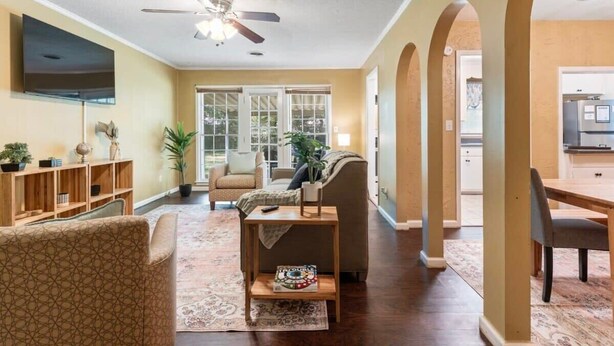 A spacious living room with a sofa, armchairs, a coffee table, and a TV mounted on the wall. There is a ceiling fan, a large window, and a door leading to the kitchen.