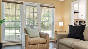 A living room with a beige sofa, a patterned armchair, and a coffee table. There are large windows with curtains, a potted plant, and a lamp on a side table.