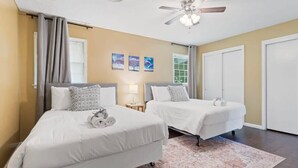 A hotel room with two beds, a ceiling fan, a window with curtains, and a wall with three framed pictures.