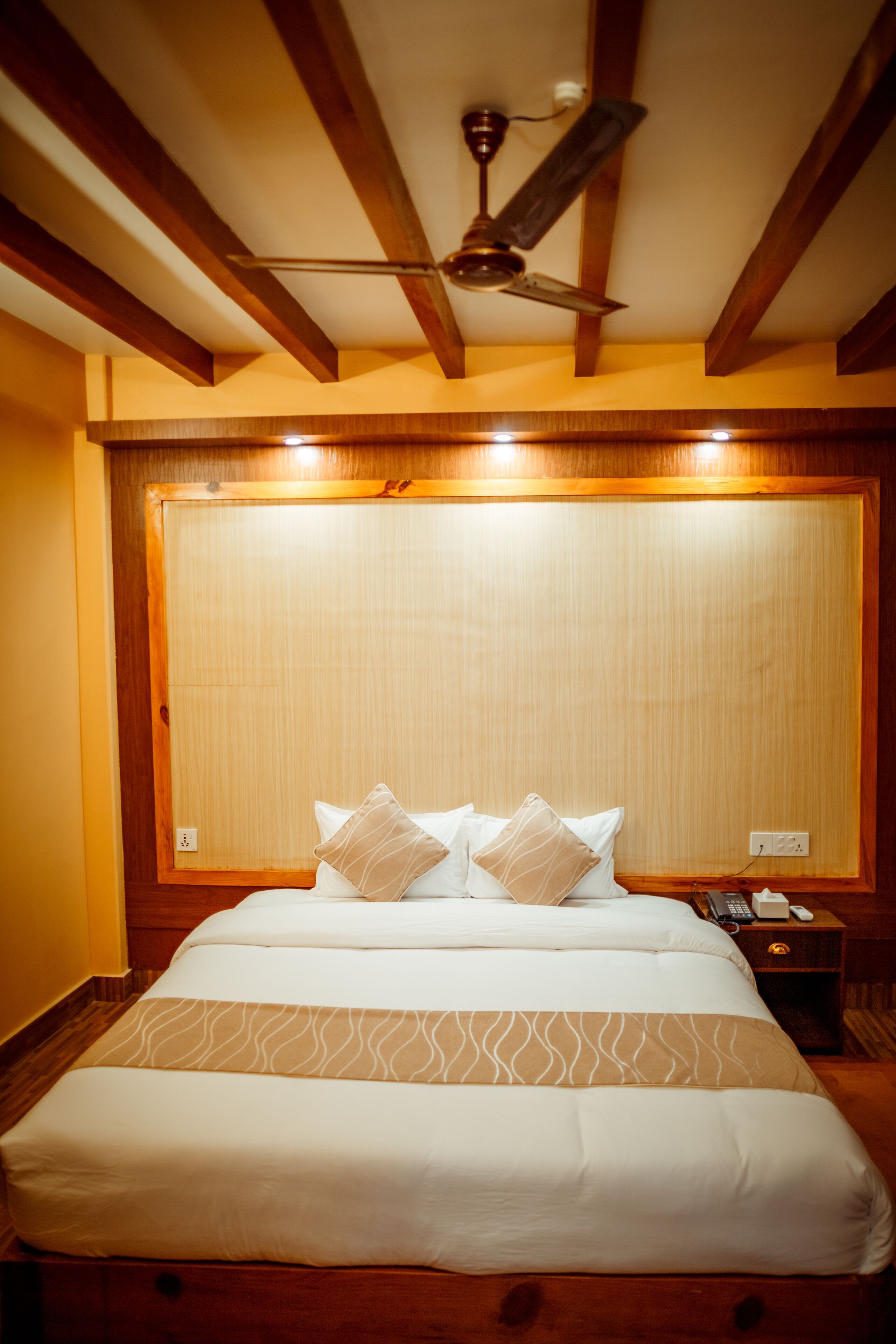 A hotel room with a large bed, wooden headboard, ceiling fan, bedside table with a phone, and a nightstand.