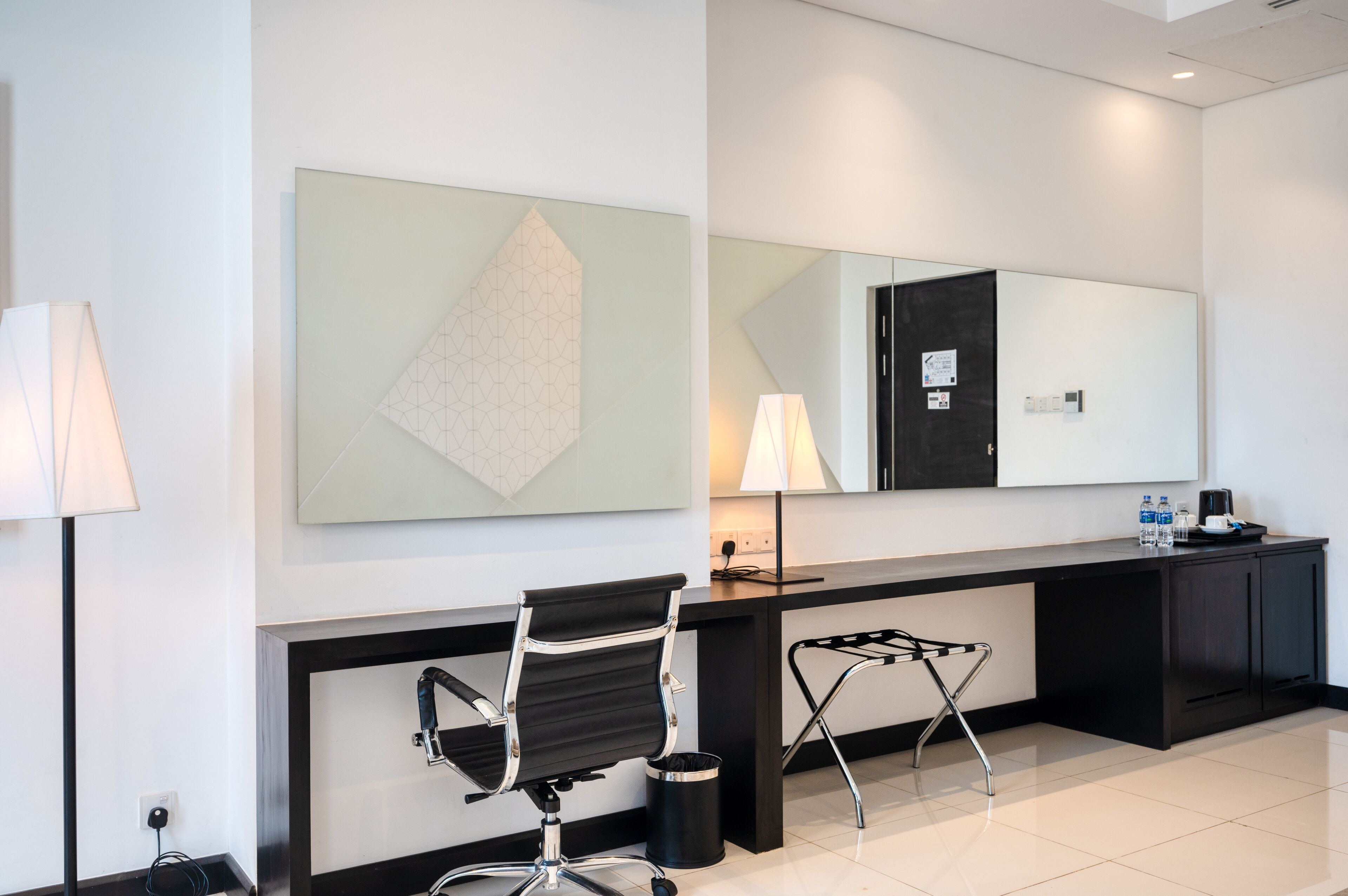 A modern hotel lobby with a reception desk, a black office chair, a standing desk, a large mirror, and a geometric artwork on the wall.
