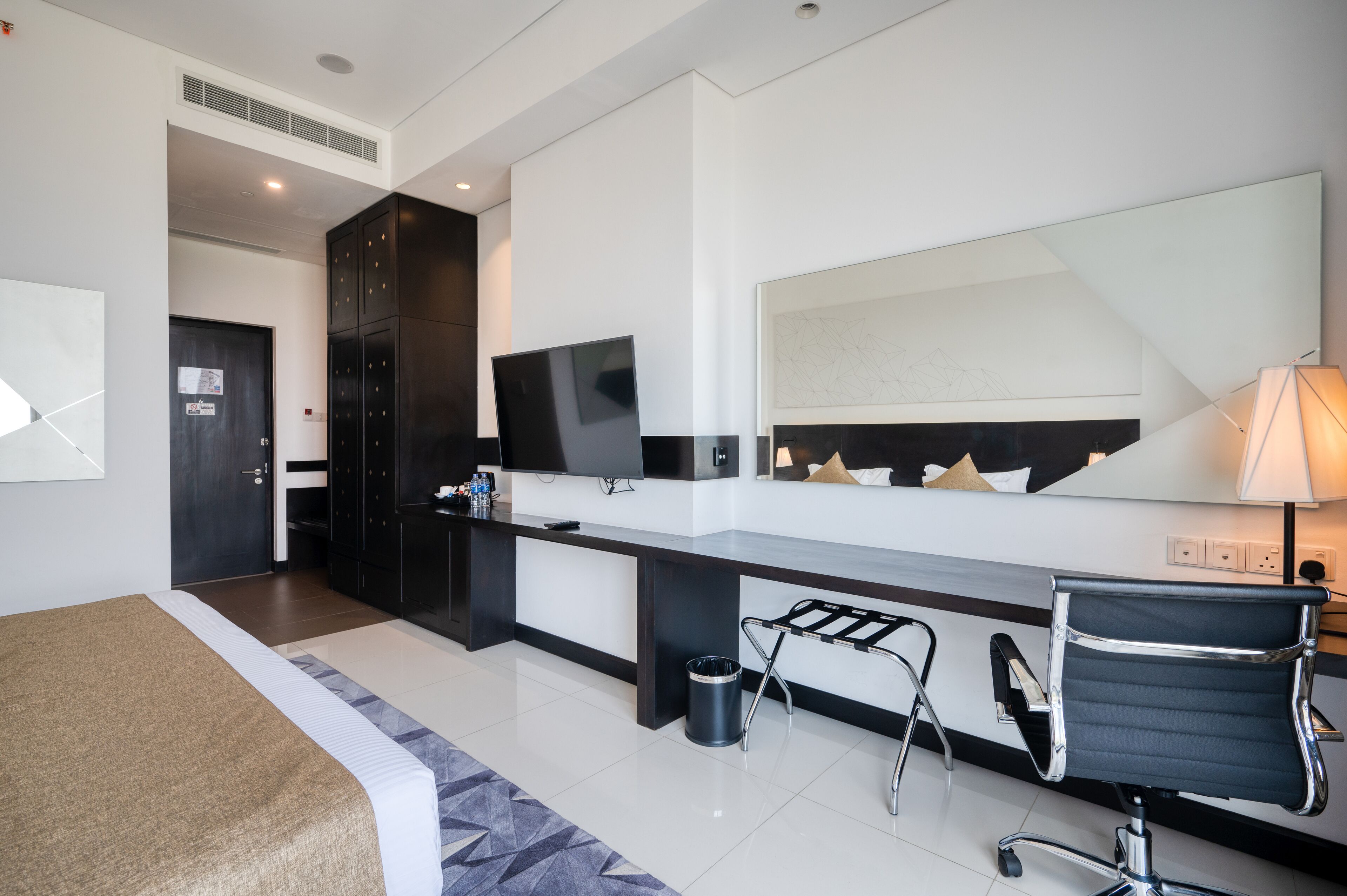 A modern hotel room with a bed, a desk, a chair, a TV, and a large mirror.