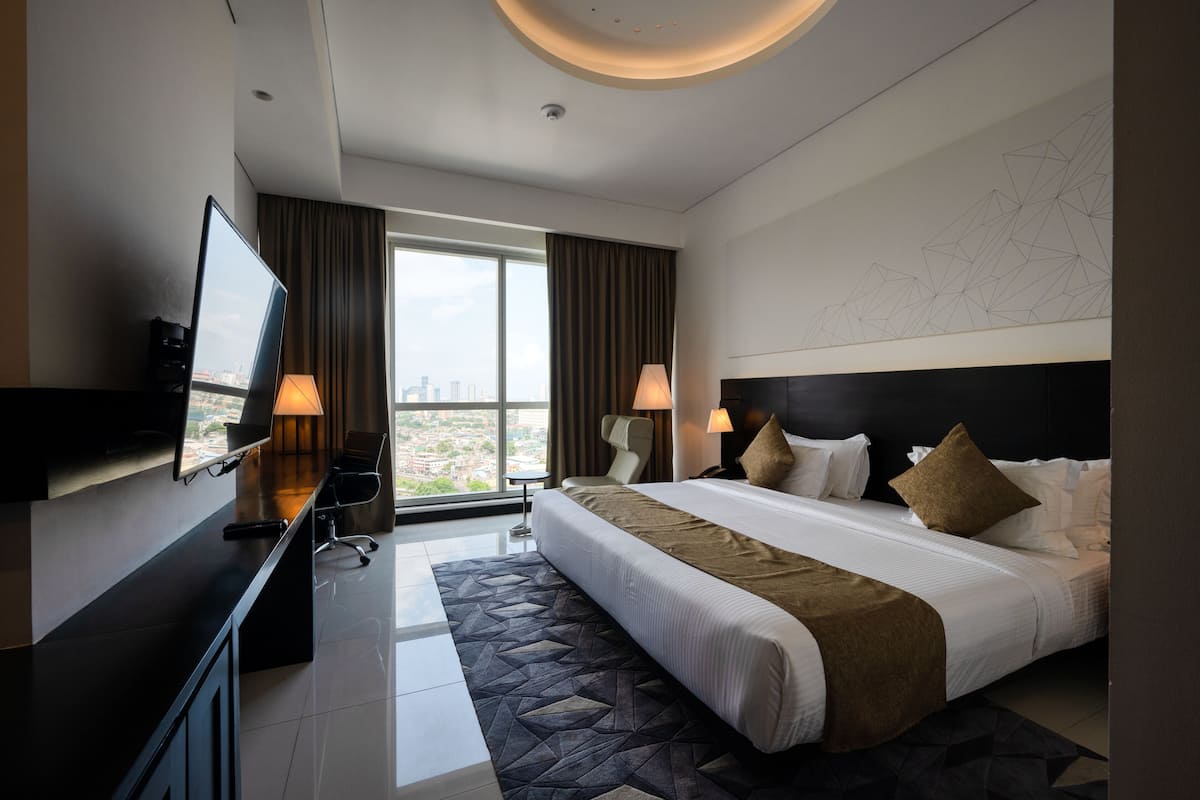 A modern hotel room with a large bed, two bedside lamps, a flat-screen TV, a desk with a chair, and a city view.