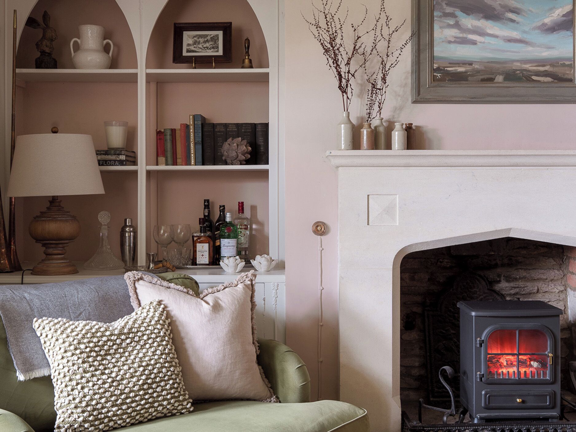 A cozy living room with a fireplace, a sofa with decorative pillows, a bookshelf with books and decorative items, and a lamp.