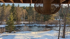 Una estructura de madera con una lámpara esférica colgante, suelo cubierto de nieve, un río y un bosque al fondo.