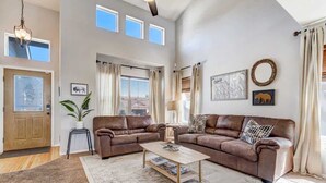 A spacious living room with a brown leather sectional sofa, a coffee table, and a large window with curtains. - Whistler Hollow! Family Home, Bball Court, Firepit (Colorado Springs)
