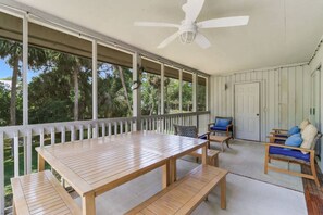 Een overdekt terras met een houten tafel en banken, een plafondventilator en uitzicht op bomen.