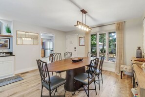 A dining area with a wooden table and chairs, a kitchen in the background, and a view of the outdoors through large windows.