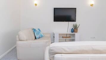Une chambre moderne avec un téléviseur à écran plat fixé au mur, un canapé blanc et un fauteuil plus petit agrémenté d’un coussin bleu.