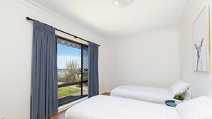 Ein Schlafzimmer mit zwei Betten, einem fenster mit Vorhängen, Blick ins Freie und einem gerahmten Bild an der Wand.
