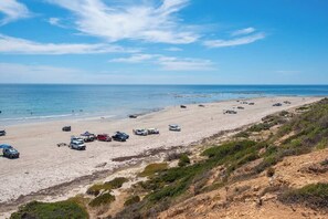 Une plage où des véhicules sont stationnés le long du rivage, un ciel dégagé et l’océan en arrière-plan.