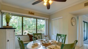 A dining area with a round table, green chairs, a ceiling fan, and a view of greenery outside.