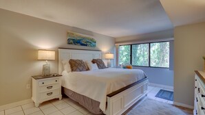 A bedroom with a large bed, bedside lamps, a nightstand, a dresser, and a window with a view of trees.