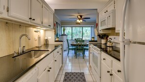 A kitchen with white cabinets, a black countertop, a sink, a ceiling fan, and a window with a view of trees.