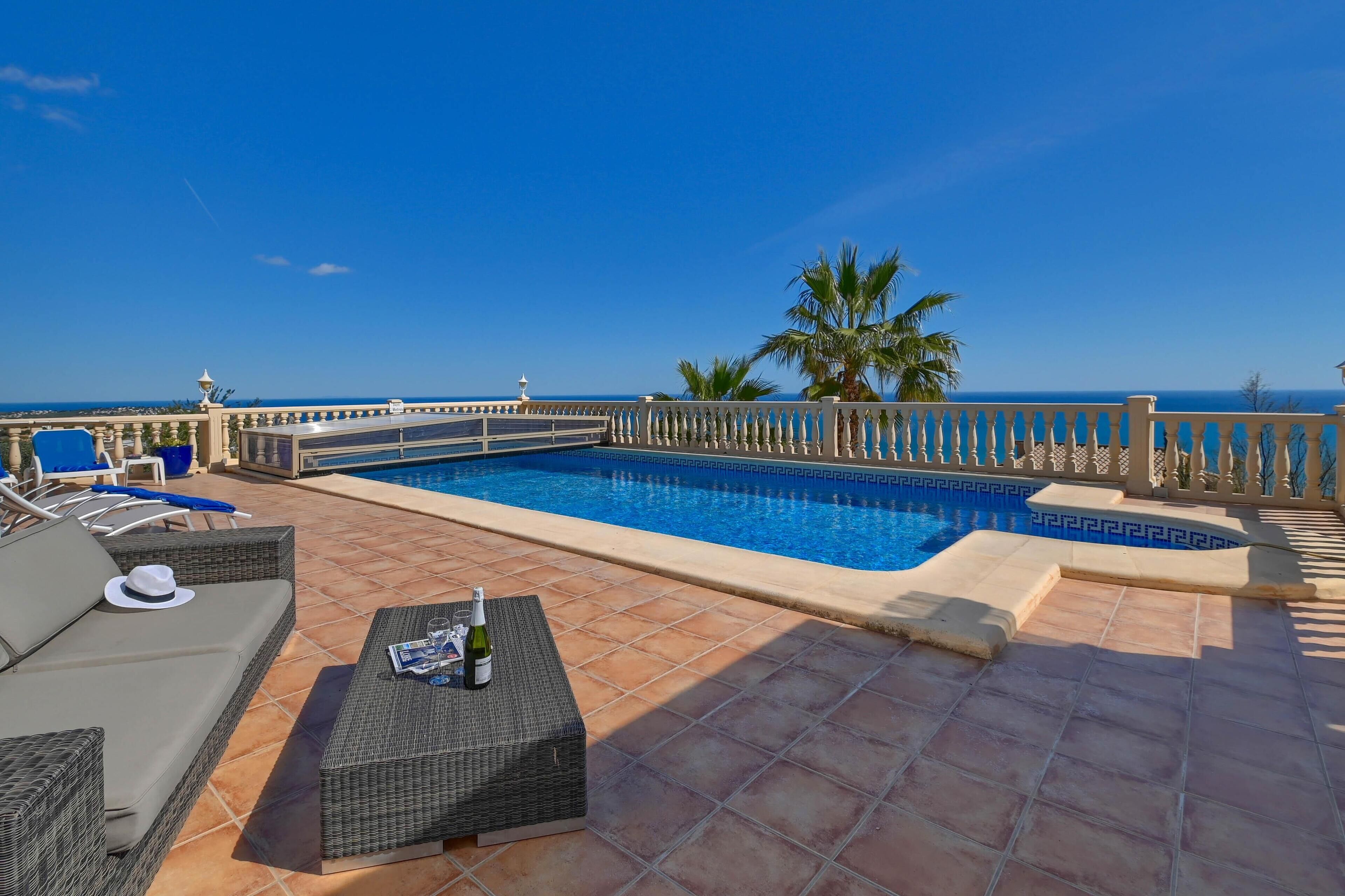 A rooftop pool area with a clear view of the ocean, a lounge chair, a wicker table with a bottle, and a tiled floor.