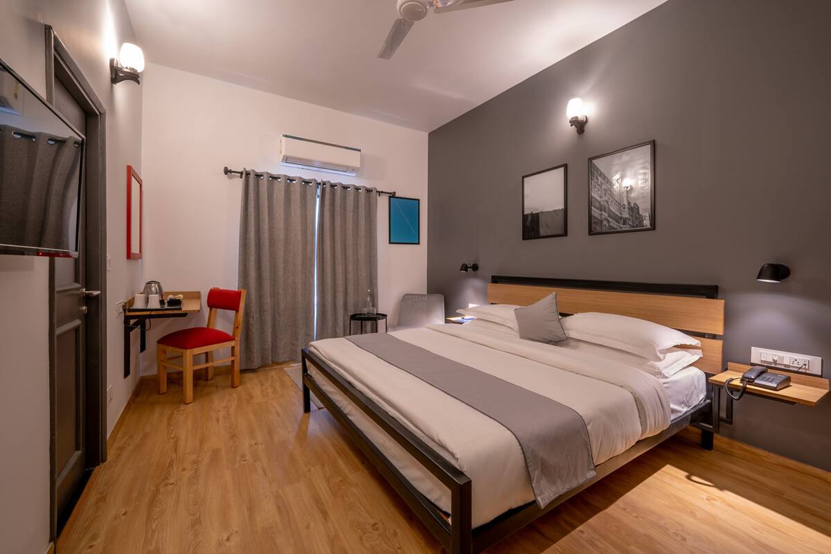 Habitación de hotel moderna con suelo de madera, cama con cabecera, escritorio con silla, televisor y ventana con cortinas.