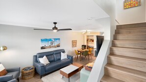 Ein modernes Wohnzimmer mit einem blauen Sofa, einem Couchtisch und einem Essbereich mit Tisch und Stühlen. Eine Treppe mit Holzboden ist sichtbar.