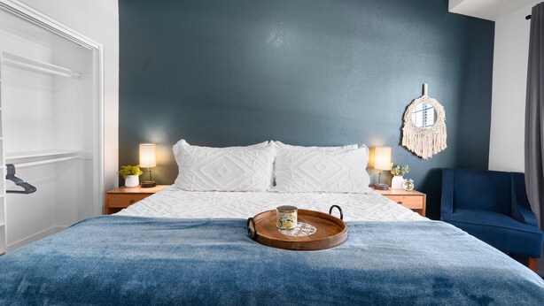 Ein Schlafzimmer mit einem Bett, Nachttischen, einem blauen Sessel, einem Holztablett mit einer Tasse und einem Wandbehang aus Makramee.