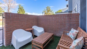 Eine Terrasse mit einer Ziegelwand, einem Holztisch, weißen Plastikstühlen und einer Holzbank mit Kissen.