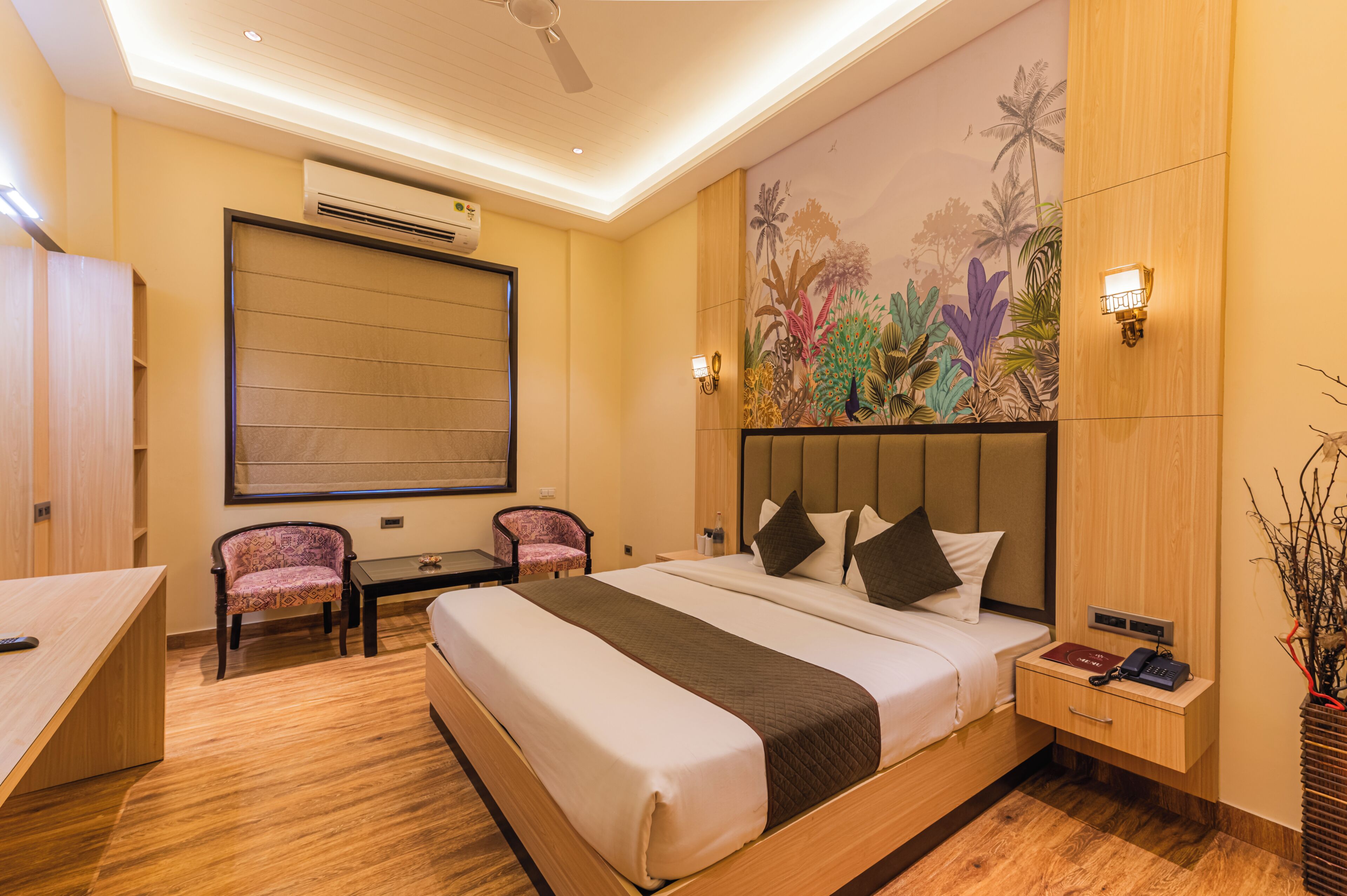 Habitación de hotel con cama, escritorio, silla, mesita de noche y un mural tropical en la pared.