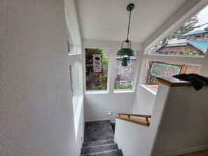 Una escalera con barandilla de madera, una lámpara colgante verde y vista de un edificio vecino a través de un ventanal.