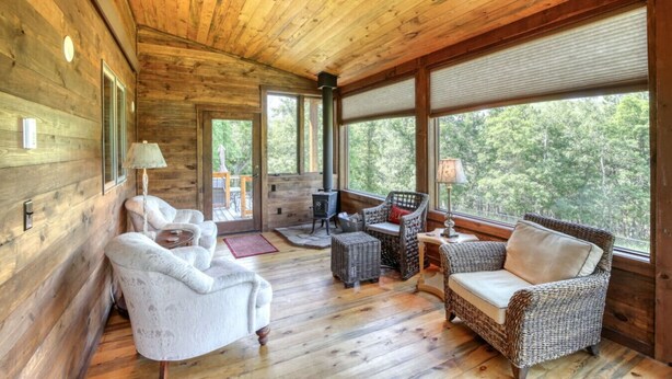 A cozy cabin interior with wooden walls, a fireplace, wicker furniture, and large windows offering a view of the forest. - Secluded & Spacious Cabin W/ a Spa and Garage (Sturgis)