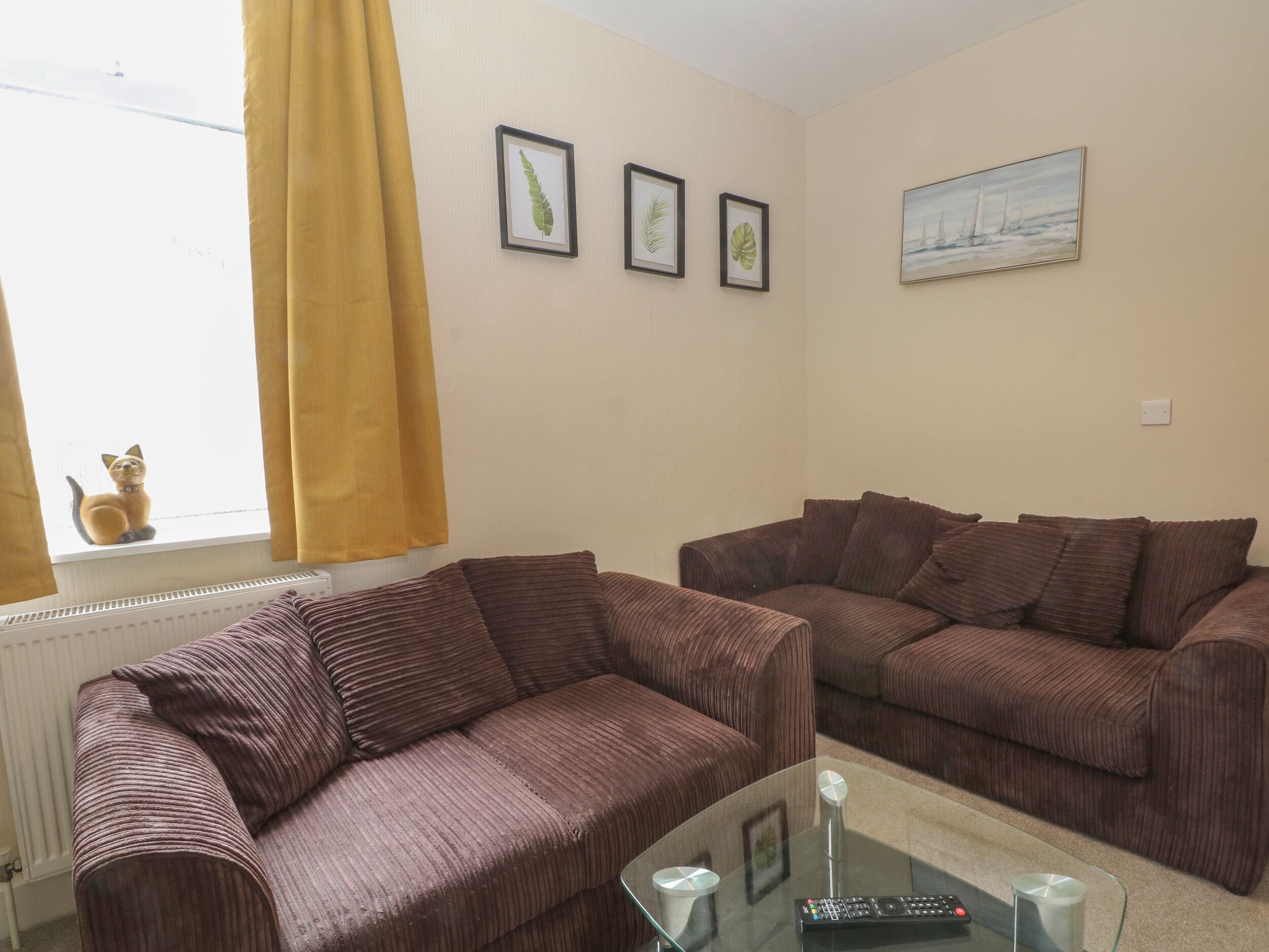 Ein Wohnzimmer mit zwei braunen Sofas, einem Glastisch und einem Fenster mit gelben Vorhängen.