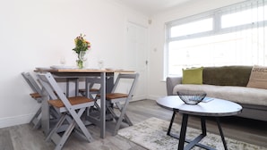 A dining area with a table and chairs, a vase with flowers, and a window with blinds. - Apple Tree Bungalow (Redruth)